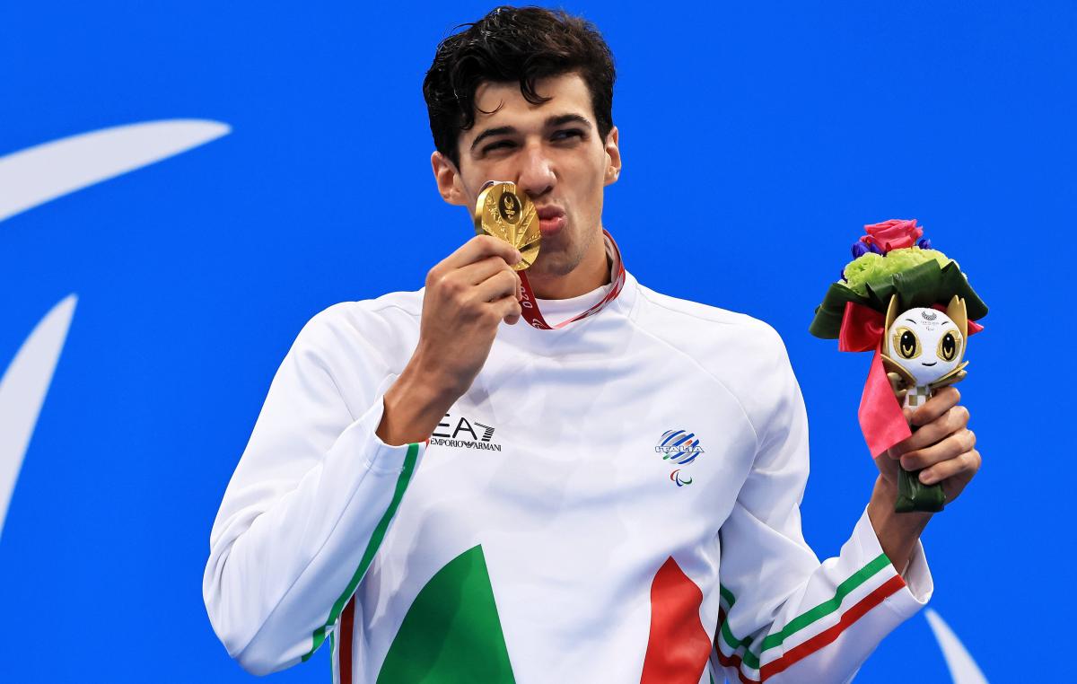 A man kisses the gold medal with a smile on his face. He is holding the miniature mascot in the other hand