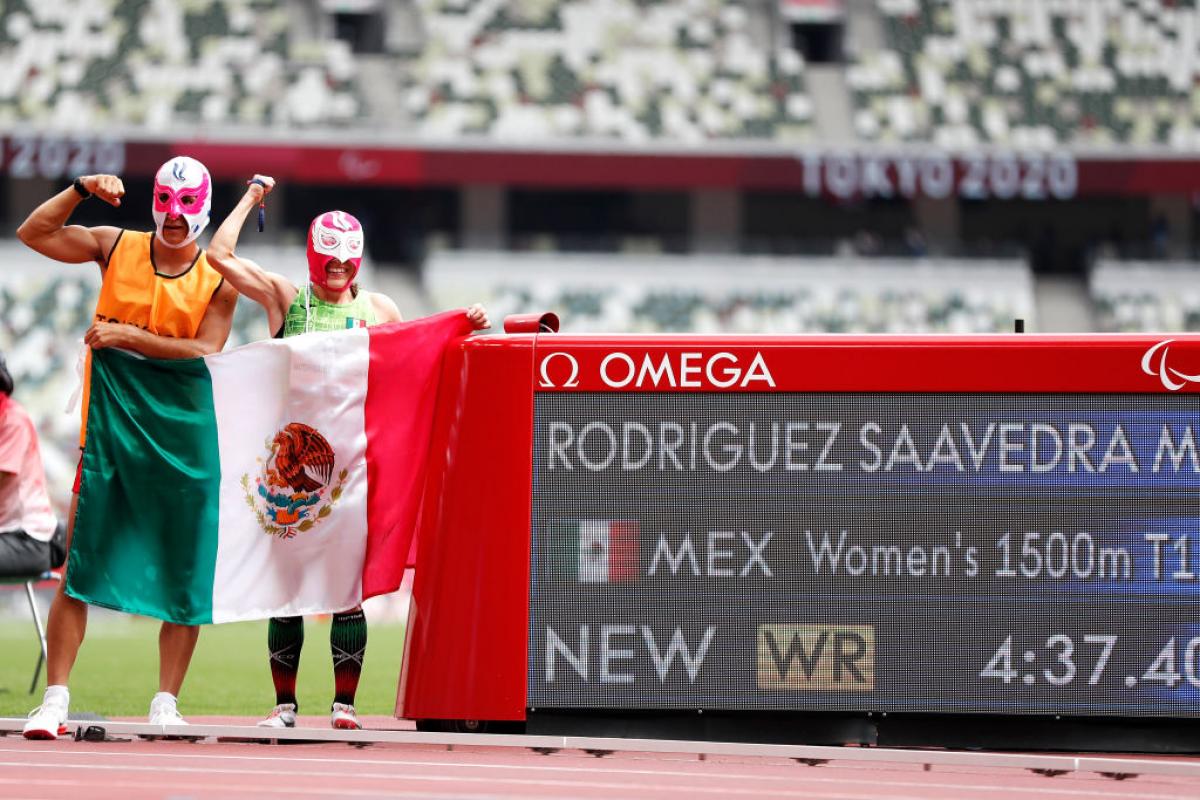 A man and a woman with Lucha Libre masks in an athletics track with the Mexican flag