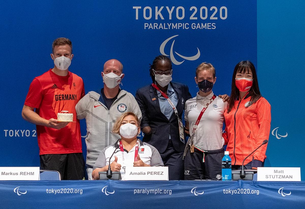 Two men and three women standing behind a seated woman in a press conference room