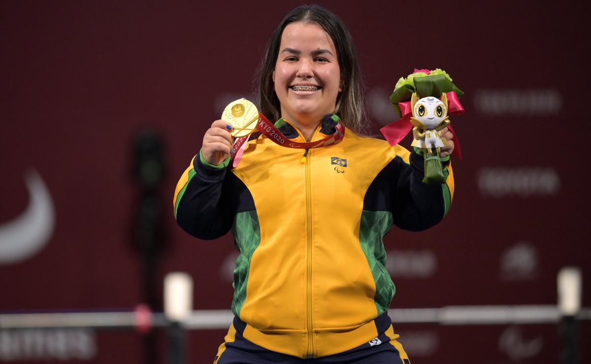 A woman posing with the gold medal in her right hand and a mascot in her left hand. She is having a big smile on her face