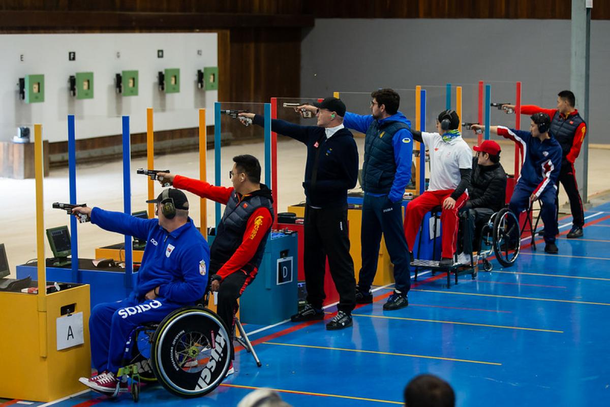 A group of eight men competing in a pistol shooting event in a shooting range