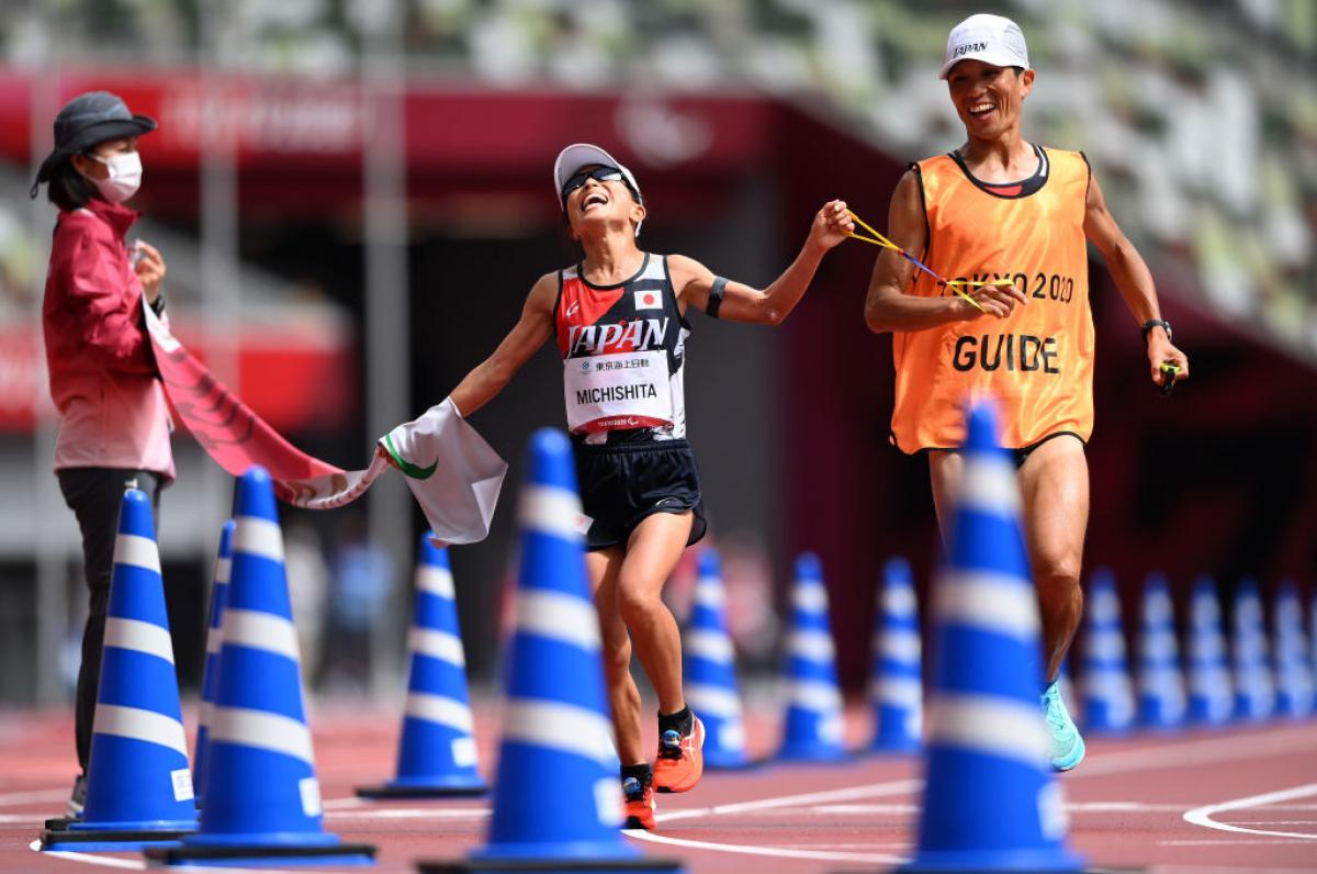 A female runner and her guide crossing the finish line in an athletics stadium