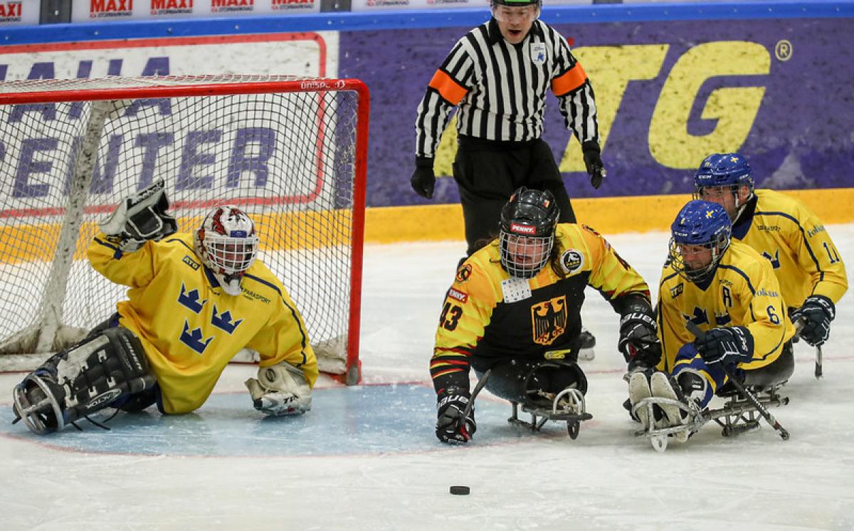 A German Para ice hockey player on ice competing against three Swedish players observed by a referee