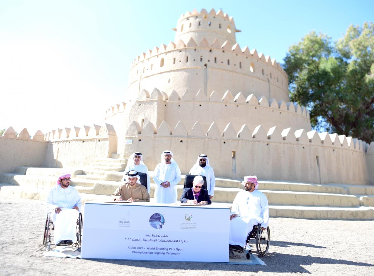 A table with a man and a woman signing a paper with two men in wheelchairs and three men standing behind them