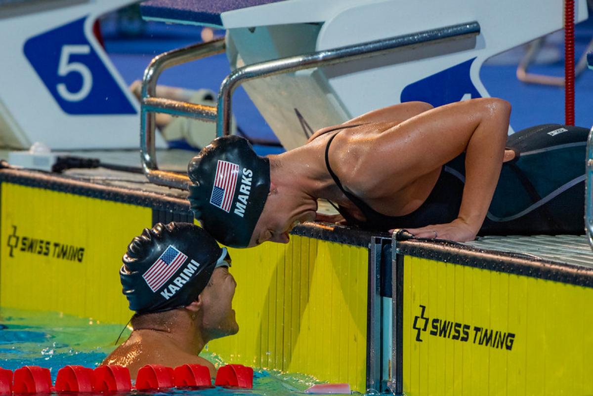 A woman outside a swimming pool stretching her head to touch the head of a man inside the pool