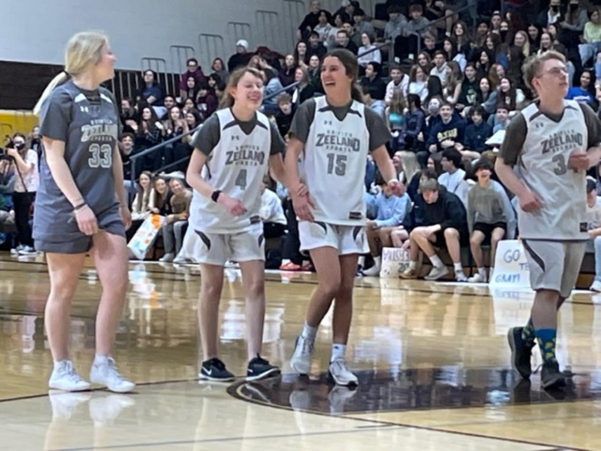 A friend guides a blind girl, both in basketball uniforms, across a basketball court in a packed stadium.