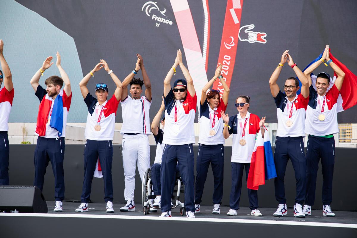 Ten people in French uniforms clap onstage with their hands raised high.