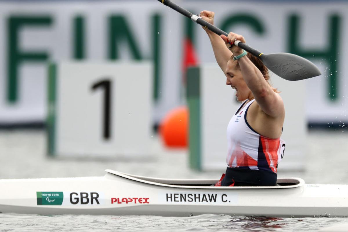 A female Para canoeist raises her paddle and yells out in celebration after crossing the finish line.