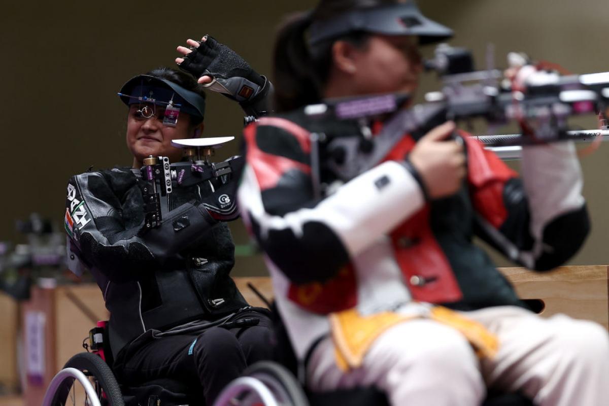 A female rifle shooter behind another shooter waving at at shooting range