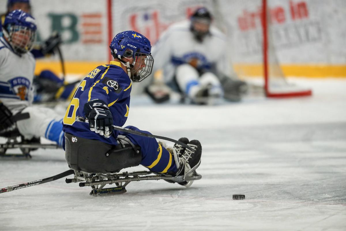 A Para hockey player preparing to shoot in front of an opponent with a goaltender in the background