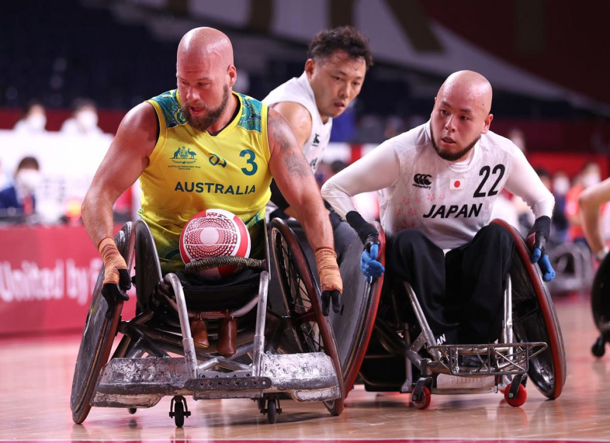 A male wheelchair rugby player in a yellow jersey carries the ball, chased by two players.