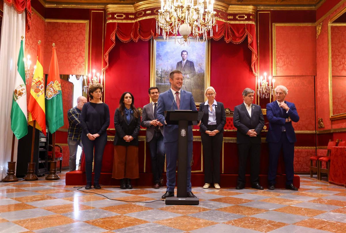 A man in a suit smiles while giving a speech at the podium in a lavishly decorated palace room.