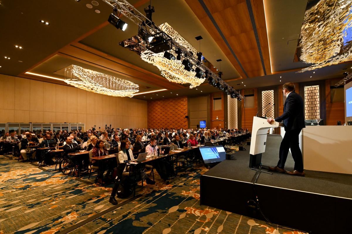 A man gives a speech from the stage as a room full of IPC member organisations looks on.