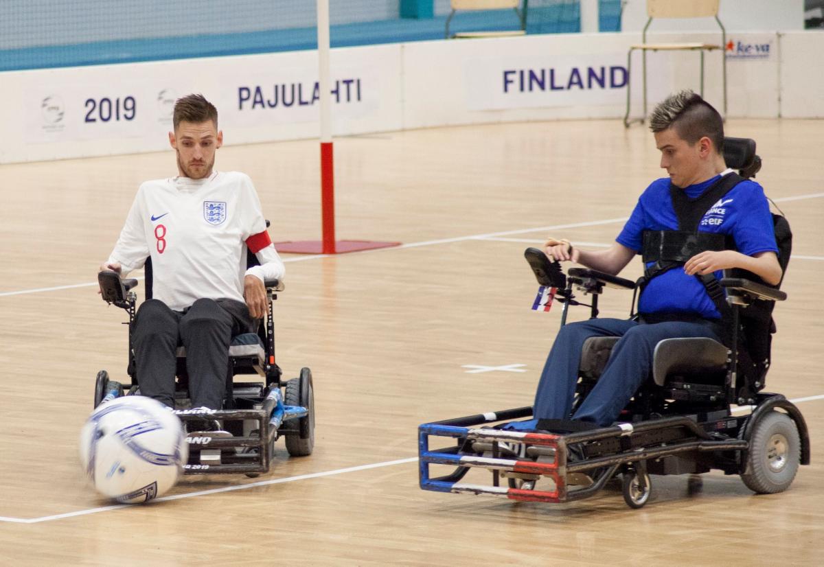 A male powerchair football athlete controls his wheelchair in front of the ball, while another player looks at the ball