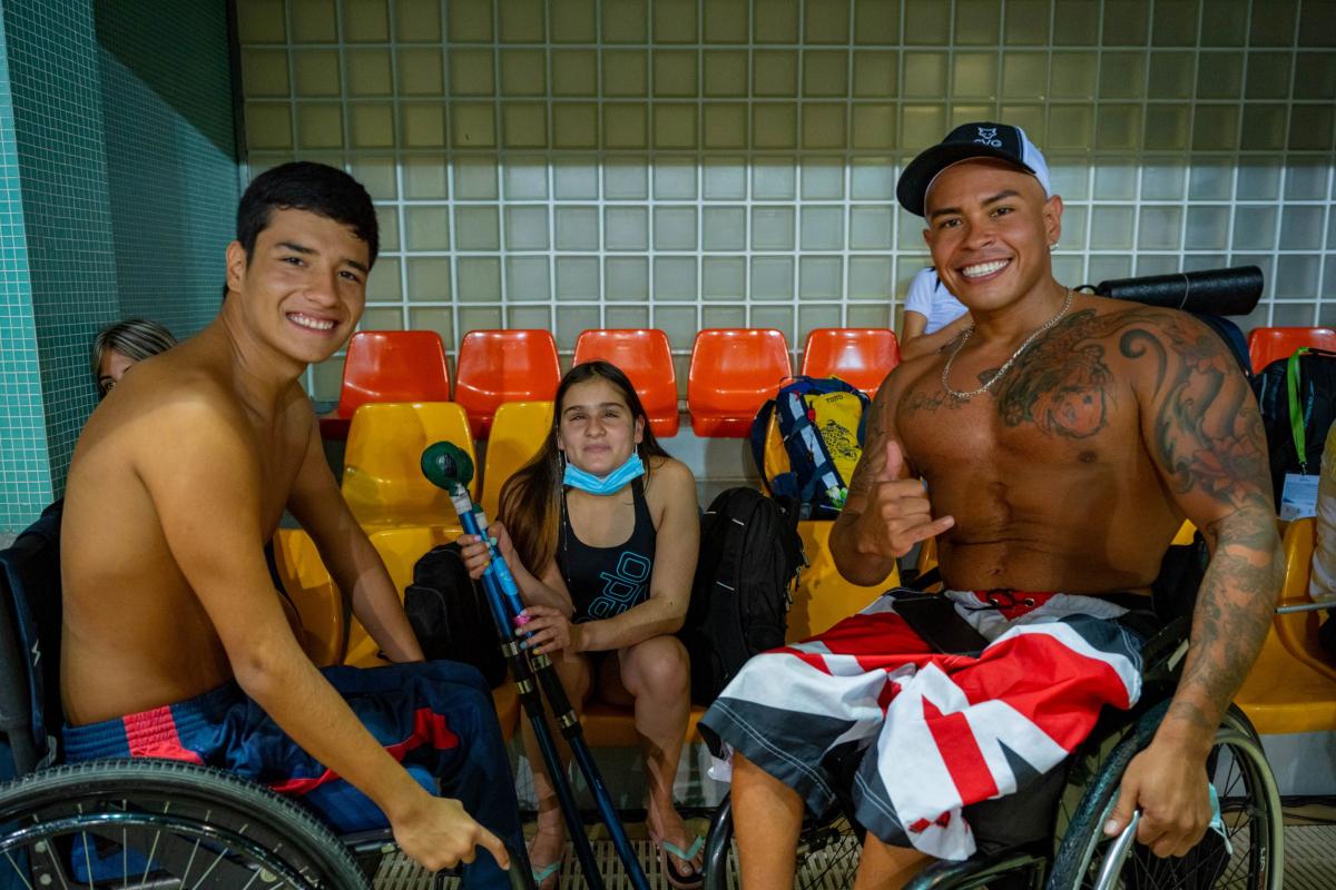 Three swimmers, two males in wheelchairs and a female swimmer holding equipment for athletes with vision impairments, pose for a photo on the bleachers.