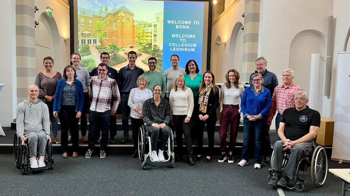 19 people pose for a photograph in front of a screen that says "Welcome to Bonn"