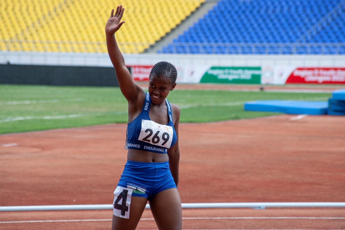 A female runner smiles and raises her right arm in a salute as she comes off the running track.