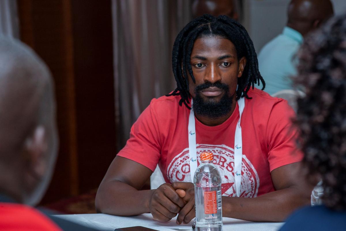 A man in a red t-shirt looks on as he sits at a table with his hands folded in front of him.