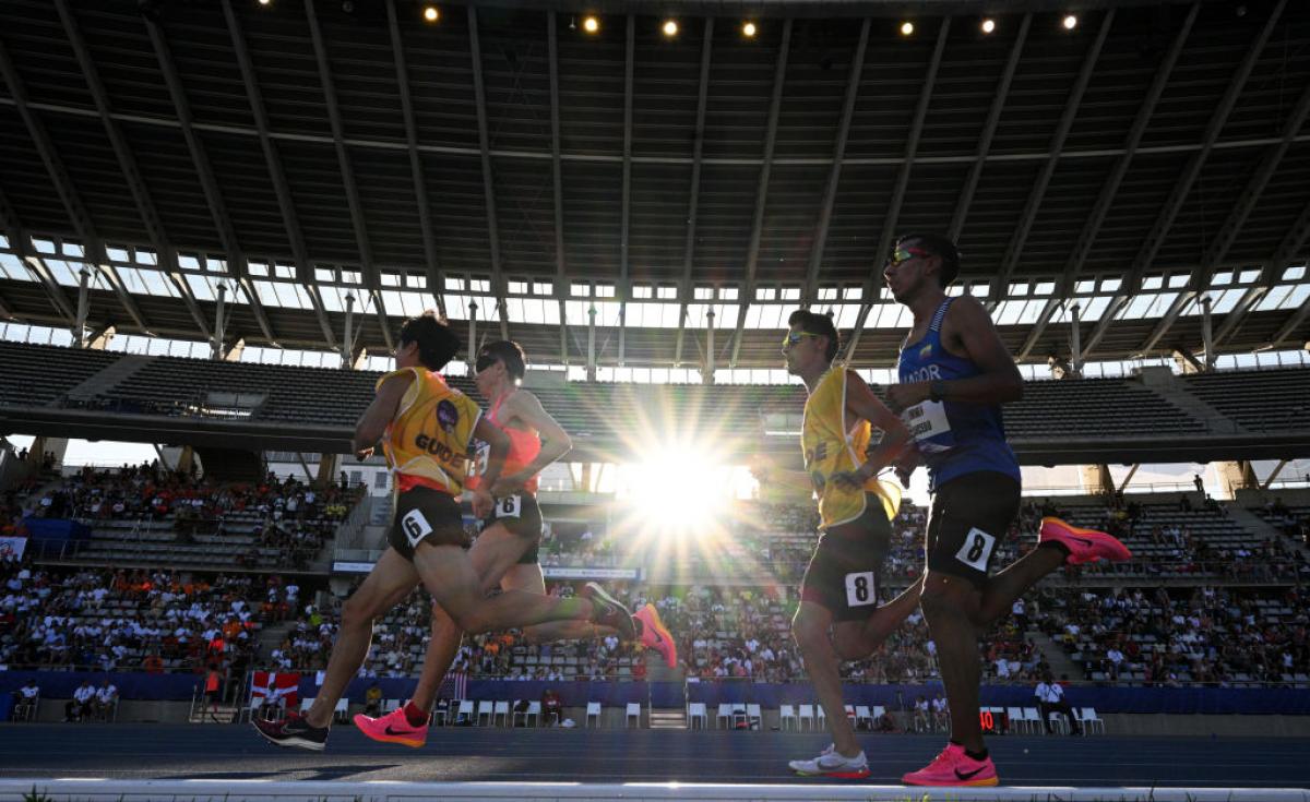 two vision impaired runners and their guides running on the track in front of spectators 