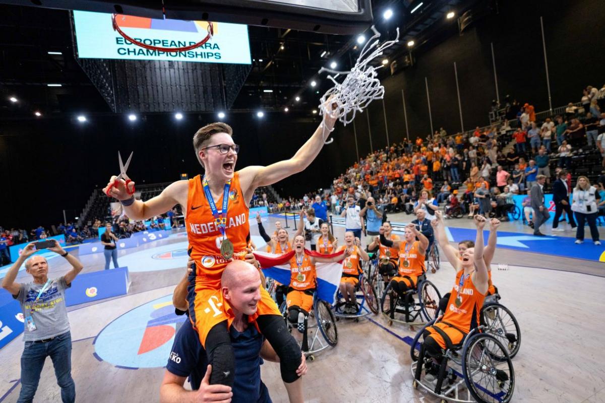 A female wheelchair basketball player cuts a basketball net, her teammates celebrate