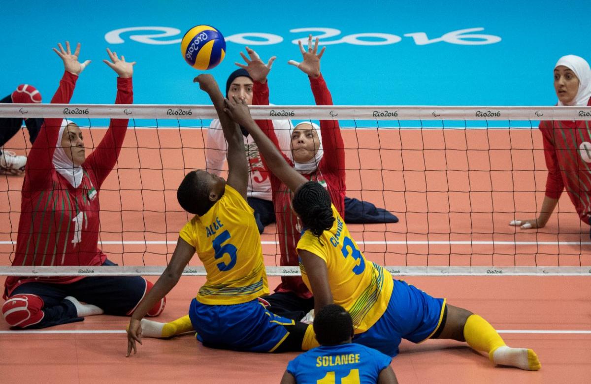 Two women's sitting volleyball teams playing at the Rio 2016 Paralympics. Two female players in yellow uniform are hitting the ball over the net, while two female players in red uniform are trying to block the ball.