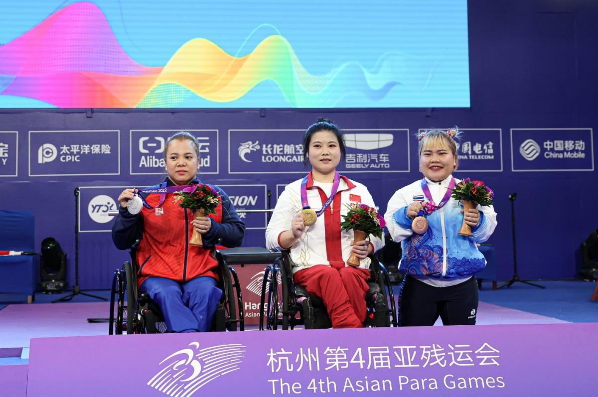 Two women in wheelchairs next to a short stature woman on a podium at the Asian Para Games