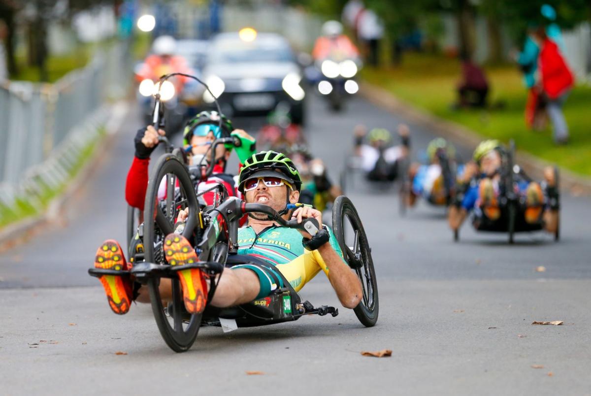 Six athletes compete on handcycles. The athlete leading the pack is wearing a South African jersey.