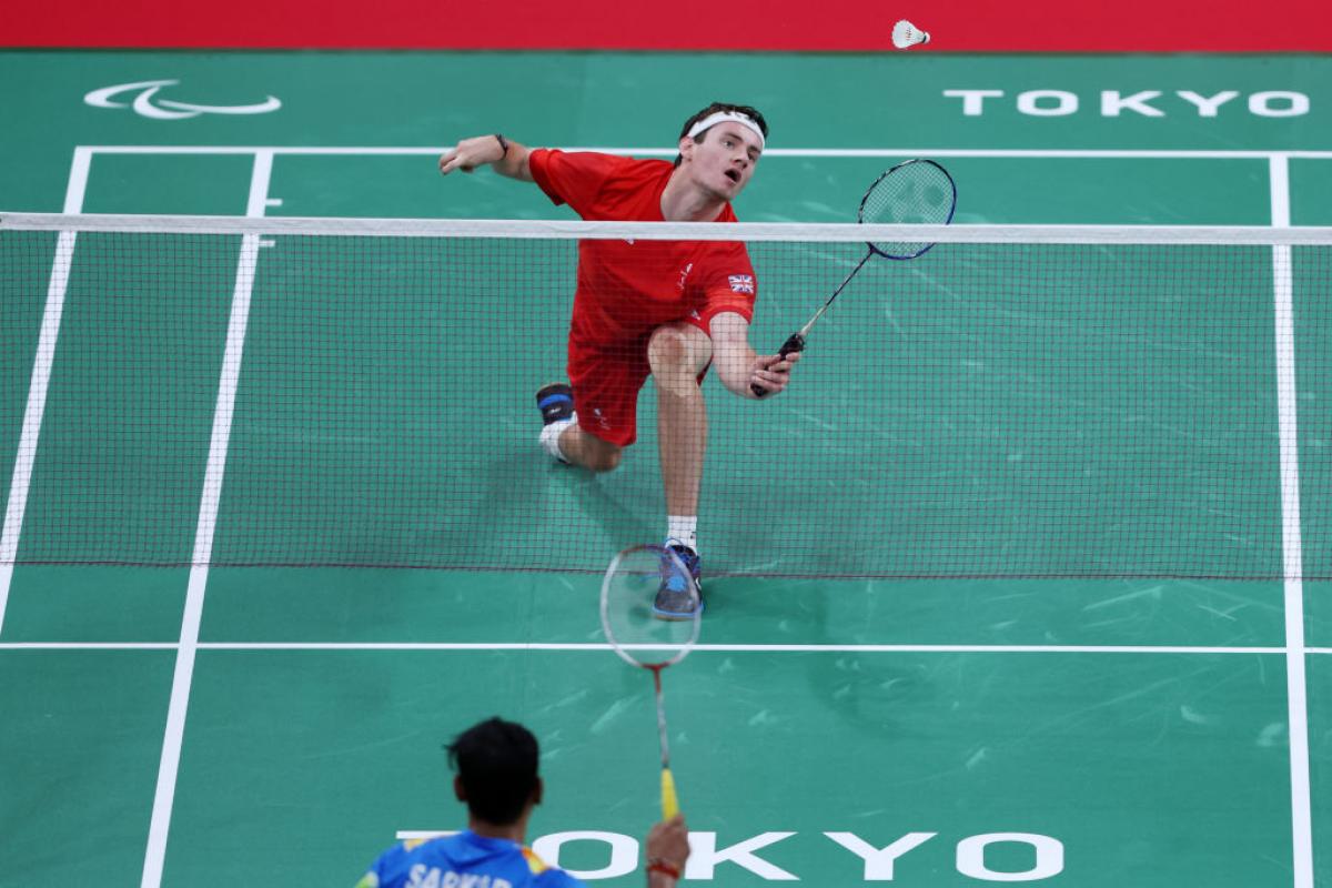 A Para badminton match between two male athletes at Tokyo 2020. A player in red uniform prepares to return the shuttle near the net.