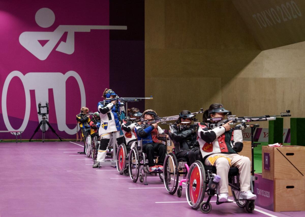 Six female athletes compete in Para shooting competition. Five athletes are competing in a wheelchair, while one athlete is standing.