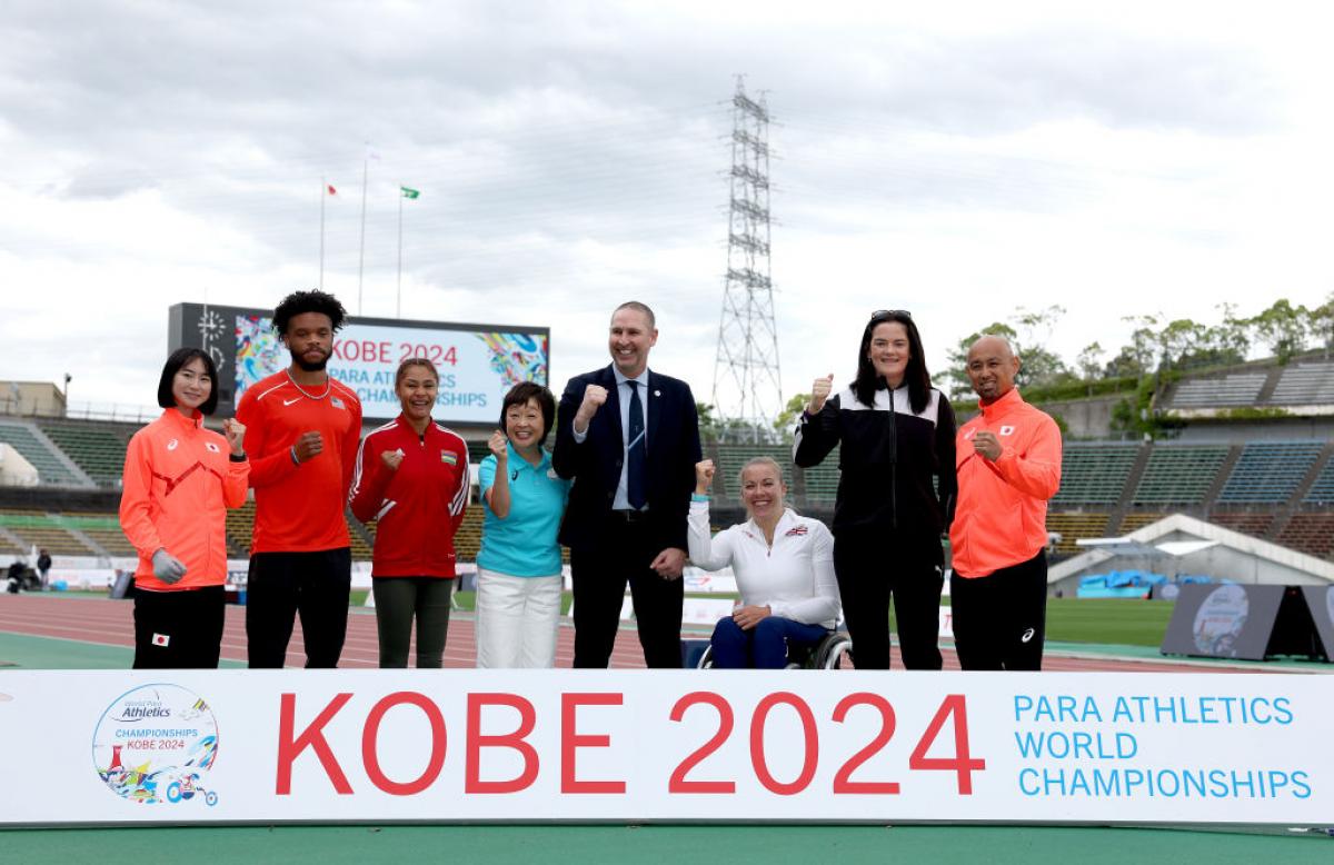 Seven standing person and a wheelchair user posing for a picture in an athletics track