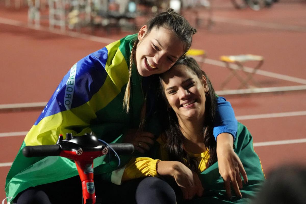 A woman in a wheelchair hugging another woman sitting on the ground