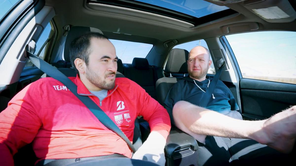 Two Para athletes are sitting in a car. One athlete is driving the car with his foot