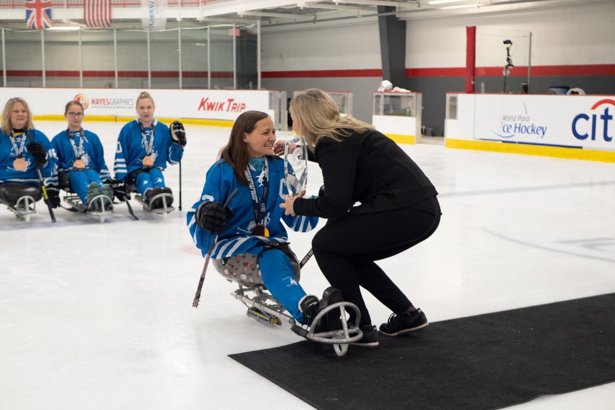 A female Para ice hockey player receiving a trophy from a woman in a hockey ring