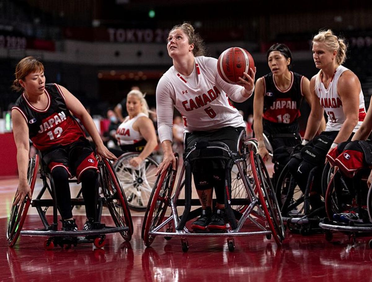 A female wheelchair basketball player carries the ball, while other players surround her