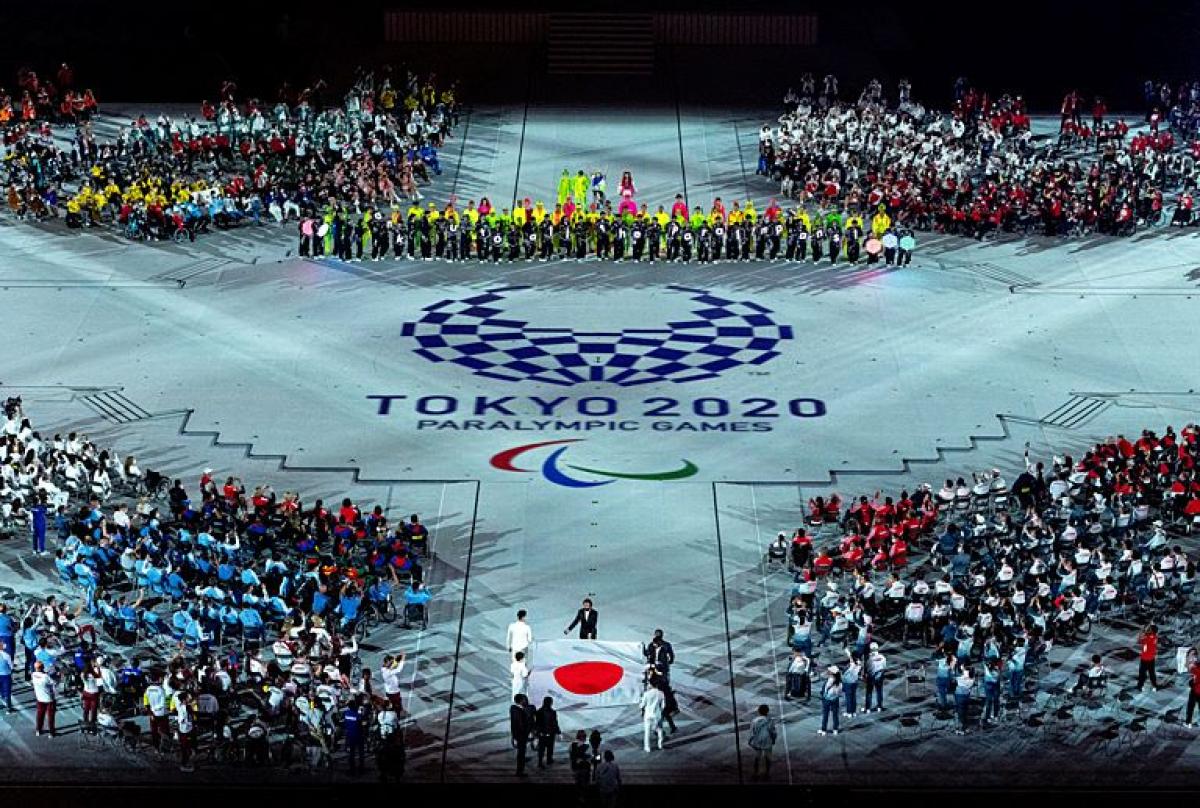 About five people carry the Japanese flag into a stadium during the Closing Ceremony of the Tokyo 2020 Paralympics. The Tokyo 2020 emblem and the Paralympic symbol is depicted on the floor.