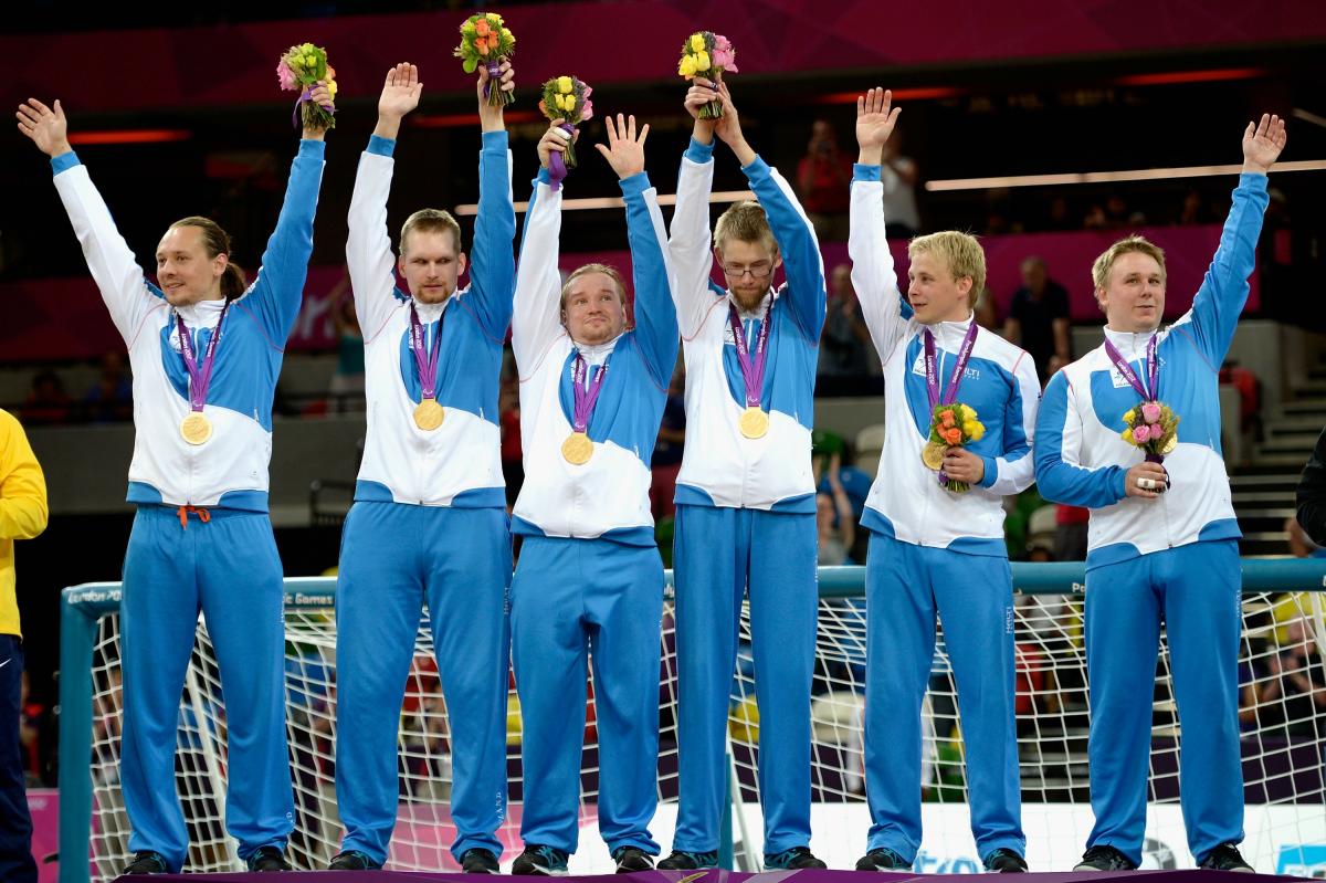 Six men in training suits standing in a row, waving and celebrating