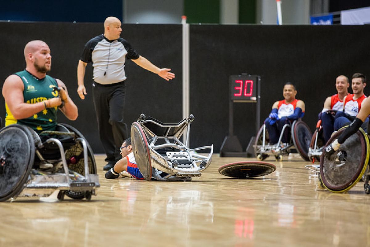 Dressed in green and seated in a wheelchair, a player looks on as another lies face down with his wheel broken from his chair.