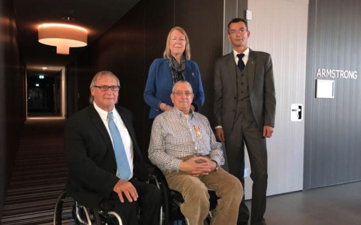 Two men in wheelchairs in suits while two people stand behind them to pose for a photo