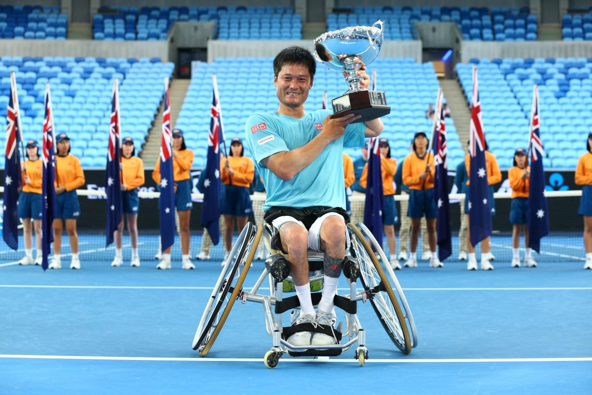 a male wheelchair tennis player lifts a trophy