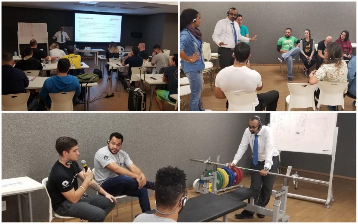 people in a room listening to a man talking and demonstrating a powerlifting bar