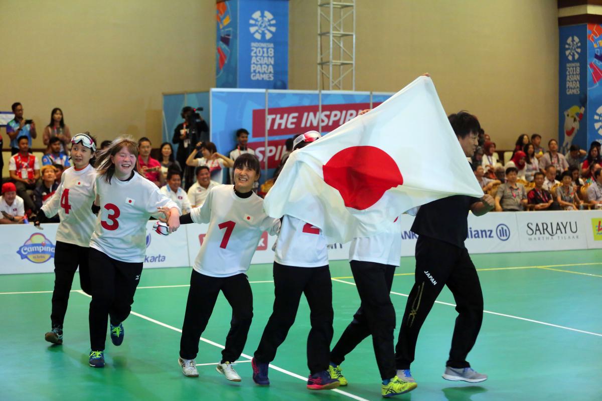 Japanese female goalball players run across the court holding a Japan  flag