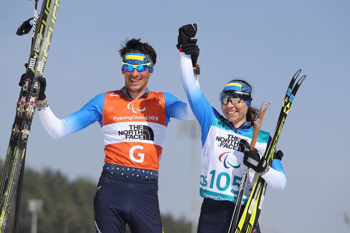 female Para Nordic skier Oksana Shyshkova holds hands with her guide as they raise their arms in victory